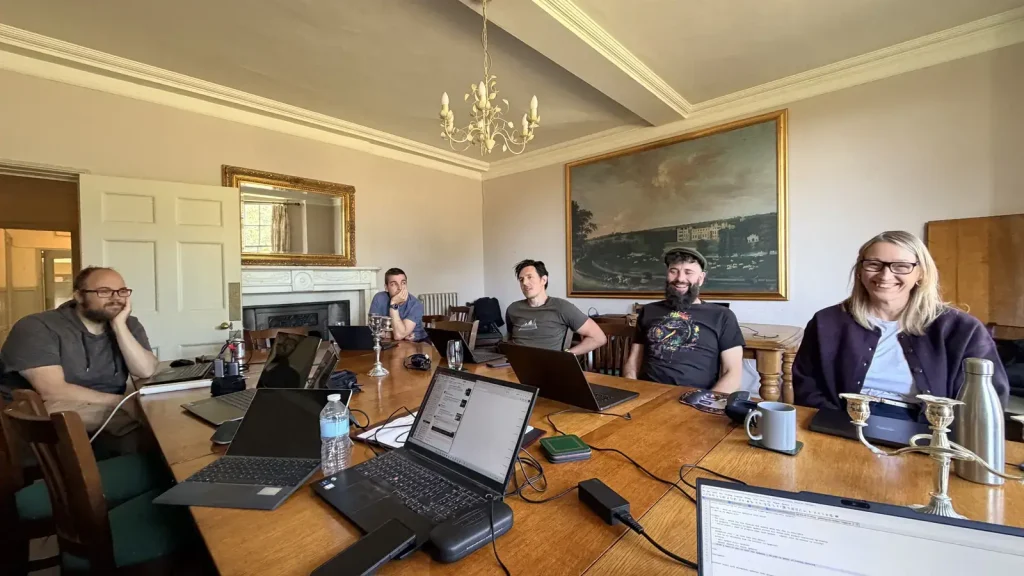 Team members sitting around a large wooden table covered in laptops, in a grand room with a chandelier and a large landscape painting.