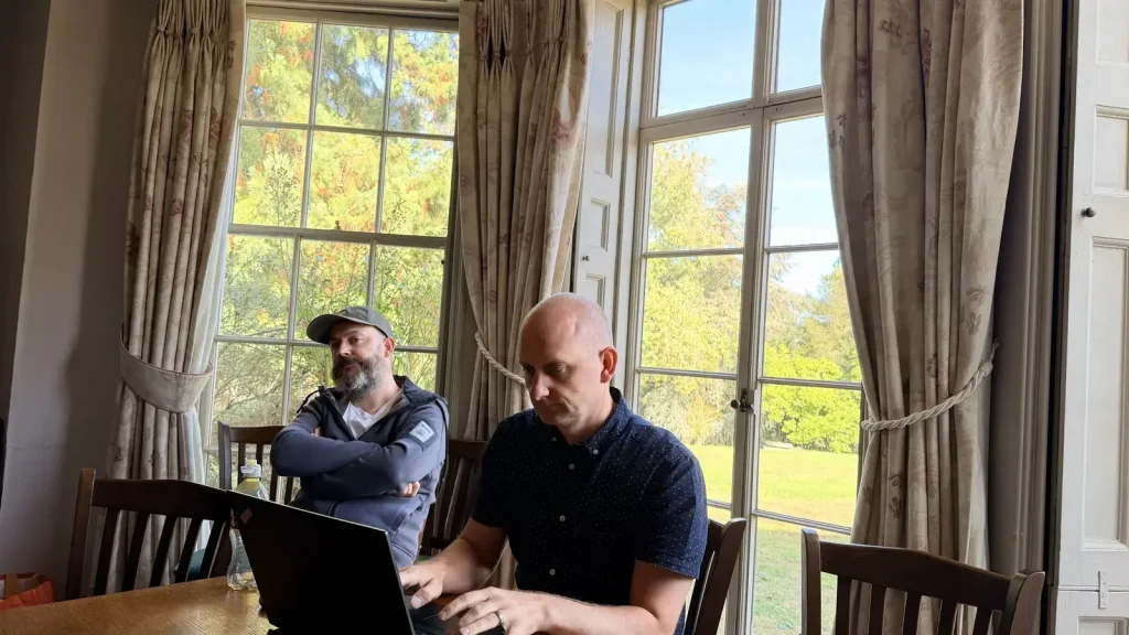 Two colleagues at a table by tall sash windows, one typing on a laptop, with sunlit gardens outside.