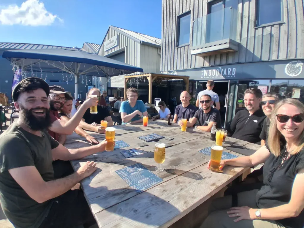 The team sitting at a long wooden table outside The Woodyard in Woodbridge, raising drinks and smiling in the sunshine.